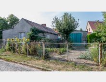 Ein kleines graues Haus mit Ziegeldach steht hinter einem Garten mit Blumen und Sonnenblumen. Der Garten ist durch einen einfachen Maschendrahtzaun mit Tor eingegrenzt, und im Hintergrund befindet sich ein Schuppen mit blauen Doppeltüren.