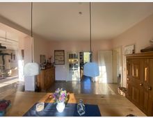 Esszimmer in einem Wohnhaus mit einem Holztisch im Vordergrund, auf dem eine Vase mit Blumen steht. Im Hintergrund sind eine Holzkommode, Glastüren mit Blick in eine Küche und ein antiker Holzschrank an einer hellen Wand zu sehen, beleuchtet durch einfallendes Tageslicht.