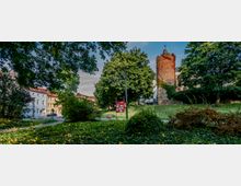 Historischer Park mit einem runden, aus Backstein gemauerten Turm im Hintergrund, umgeben von Grünflächen, Bäumen und einzelnen Sträuchern; links stehen mehrstöckige Wohnhäuser mit roten Dächern.