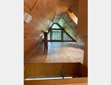 Dachgeschossraum mit schrägen Holzverkleidungen an Wänden und Decke, Holzdielenboden und einem dreieckigen Fenster mit Balustrade im Hintergrund. Eine offene Bodenluke im Vordergrund führt in den unteren Bereich.