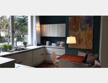 Moderne Küche mit weißen Hochglanzmöbeln, einer großen Fensterfront mit Blick auf einen Garten, und einer Eckbank mit Holztisch. An der dunkelgrünen Wand hängt ein großes, abstraktes Kunstwerk, darüber eine schlichte Deckenleuchte.