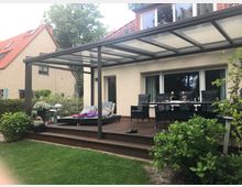 Überdachte Holzterrasse an einem Wohnhaus mit großem Glasdach und Metallstützen, davor Rasen und Gartensträucher. Unter der Überdachung stehen Liege, Sitzgruppe und Esstisch vor einer breiten Schiebetür zur Hausfassade.