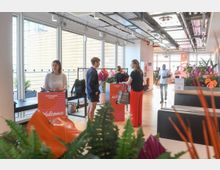Bright modern office lobby with large floor-to-ceiling windows, exposed ceiling pipes and cable trays, and light wood flooring. Welcome check-in counters and coat racks sit among indoor plants and lounge seating along a wide corridor.