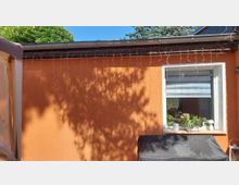 Außenwand eines eingeschossigen Hauses mit orangefarbenem Putz, Dachrinne und einer Lichterkette entlang der Traufe. Rechts ein weiß gerahmtes Fenster mit Fensterbank, auf der Pflanzen und kleine Dekoration stehen; darunter ein abgedeckter Grill, im Hintergrund Baumschatten.