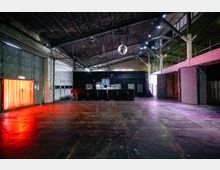 Großer, leerer Industriebau mit hohen Decken, sichtbarer Metallkonstruktion und einer glänzenden Discokugel in der Mitte. An der Rückwand befindet sich eine schwarze Bar mit hellen Getränkekühlschränken, während bunte Lichter eine rote und violette Stimmung erzeugen.