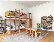Helles Kinderzimmer mit hohen weißen Wänden und Holzfußboden, ausgestattet mit einem großen Etagenbett aus Holz mit Leiter, Stauraum und Hängeseil. In der Raummitte steht ein kleiner Tisch mit Stühlen auf einem grauen Teppich; an den Wänden stehen Regale und Schränke mit Büchern und Spielzeug.
