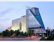 Modernes Hotelgebäude mit einer auffälligen Architektur, bestehend aus weißen Fassaden und großen Glasflächen in einer schrägen, prismatischen Struktur. Im Vordergrund sind Bäume, eine Straße mit Lichtspuren und mehrere Fahnenmasten sichtbar.