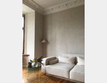 Helles Wohnzimmer mit beigefarbener Couch, einem gläsernen Beistelltisch mit Blumenarrangement, stehendem Lampenschirm und einer grau verputzten Wand. Der Raum hat eine dekorative Stuckdecke und einen Holzboden.