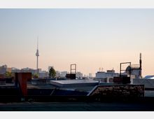 Blick über Flachdächer mit Schornsteinen und technischen Anlagen, im Hintergrund die Skyline von Berlin mit dem Fernsehturm bei klarem Himmel.