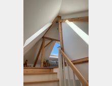 Blick vom Treppenaufgang in ein helles Dachgeschoss mit schrägen weißen Wänden und sichtbaren Holzbalken. Ein großes Dachfenster lässt Sonnenlicht auf eine kleine Sitzecke mit Sofa und Beistelltisch fallen.
