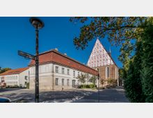 Straßenecke mit einer weiß verputzten, zweigeschossigen Gebäudereihe mit roten Ziegeldächern, daneben eine große Backsteinkirche mit steilem, weiß-rot gemustertem Giebel und hohen Spitzbogenfenstern. Im Vordergrund stehen ein Laternenmast mit Straßenschild sowie Bäume und Grünbewuchs am Straßenrand.