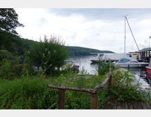 Ufer eines Sees mit dichtem Grün am Vordergrund, daneben ein Holzsteg. Im Hintergrund liegen mehrere Boote und ein Ausflugsschiff am Wasser, umgeben von bewaldeten Hügeln und einer bewölkten Himmel.