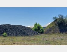 Ein offenes Gelände mit mehreren Hügeln aus dunklem Schotter, dazwischen einzelne Büsche und Grasflächen. Im Hintergrund ragen weiße, zeltartige Strukturen über die Hügel hinaus, der Himmel ist klar und blau.