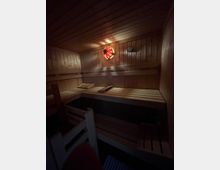 Eine Sauna mit Holzwänden, -decken und -bänken in mehreren Ebenen. Im Hintergrund ist eine Leuchte in Rottönen und ein Thermometer sichtbar, die Beleuchtung ist gedämpft.