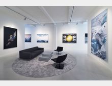 Moderne Kunstgalerie mit weiß gestrichenen Wänden, mehreren großformatigen Gemälden und Fotografien. Im Vordergrund stehen ein graues Sofa, zwei schwarze Stühle und ein niedriger rechteckiger Tisch auf einem runden Teppich.