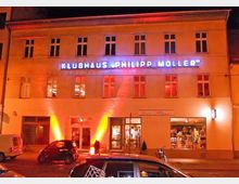 Beleuchtetes Gebäude bei Nacht mit der Aufschrift „Klubhaus Philipp Möller“ in blauen Leuchtbuchstaben. Unten befinden sich große Fenster und Geschäftseingänge, teilweise rot angestrahlt, während Autos vor dem Gebäude parken.