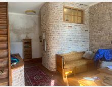 Wohnraum mit rustikalem Design, geprägt von unverputzten Ziegelwänden und Holzmöbeln. Im Vordergrund ein gelbes Sofa vor einer halbrunden Backsteinwand mit eingelassenem Fenster, umgeben von einem roten Teppich, einer Holztreppe und Regalen.