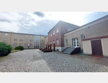 Ein gepflasterter Innenhof mit einem roten Backsteingebäude und einem beige verputzten Gebäude. Links im Hintergrund befindet sich ein weiteres mehrstöckiges Gebäude mit schmalen Fenstern und einer Glasfassade in der Mitte. Im Vordergrund steht eine Metallskulptur mit der Aufschrift „FREI“.