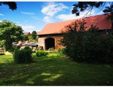 Backsteinbau mit rotem Ziegeldach in einem grünen Garten, umgeben von Büschen, Bäumen und einer Rasenfläche; im Hintergrund sind weitere Gebäude und handwerkliche Materialien zu sehen. Der Himmel ist blau mit vereinzelten Wolken.