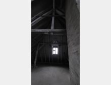 Dunkler Dachboden mit sichtbaren Holzbalken und einer kleinen Fensteröffnung, die schwaches Tageslicht in den Raum lässt. Die Wände bestehen aus unverkleidetem Material, und der Boden scheint aus Beton zu sein.