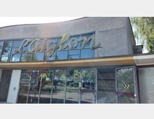 Eingangsbereich eines verlassenen Gebäudes mit dem Schriftzug „L’Aiglon“ in geschwungener Leuchtschrift über großen Glasfenstern. Die Scheiben sind teilweise mit Graffiti bemalt, und die Umgebung spiegelt sich darin.