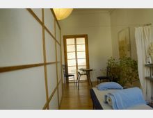 Behandlungsraum mit minimalistischer Einrichtung, bestehend aus einer Massageliege mit blauen Handtüchern, einem runden Glastisch mit Stühlen und einer Trennwand aus Holz und Papier. Im Hintergrund befinden sich eine Glasschiebetür, ein Vorhang sowie eine Zimmerpflanze.