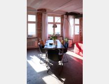 Ein Wohnzimmer mit industriellem Stil, erkennbar an den unverputzten Backsteinwänden und freiliegenden Stahlträgern. Im Vordergrund steht ein Esstisch mit schwarzen Stühlen, dekoriert mit Blumen, hinter einem großen Fenster mit weißen Vorhängen.