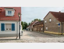 Gepflasterte Straße in einer Wohngegend mit niedrigen Häusern, teils moderner, teils älterer Bauart. Im Vordergrund ein Haus mit roten Ziegeldach und blauen Fensterläden, daneben Laternen und Straßenschilder.