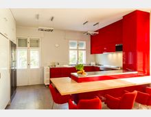 Moderne Küche mit roten Hochglanzschränken, einer zentralen Kücheninsel mit heller Arbeitsplatte und roten Stühlen. Im Hintergrund befinden sich weiße Wände, Fenster mit Rollläden und ein Kühlschrank auf der linken Seite.