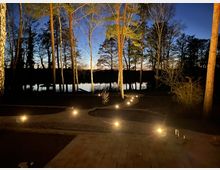 Eine beleuchtete Terrasse im Vordergrund, umgeben von hohen Bäumen, mit Blick auf einen ruhigen See im Hintergrund; die Abenddämmerung zeichnet den Himmel in dunklen Blautönen. Wege mit Bodenspot-Leuchten führen durch den Gartenbereich zur Uferpromenade.