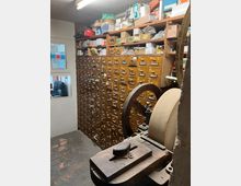 Ein kleiner Raum mit einer Wand voller hölzerner Schubladenschränke mit Beschriftungen, die zahlreiche kleine Fächer umfassen. Im Vordergrund steht eine alte mechanische Maschine mit einer großen Schleifscheibe, und auf den Schränken liegen diverse Kisten und Materialien.