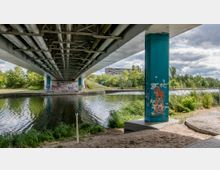 Ansicht unter einer Brücke mit einem Fluss, umgeben von Bäumen und Grünflächen; eine Brückenstütze mit farbigen Graffiti im Vordergrund und ein gepflasterter Weg daneben. Im Hintergrund ist ein mehrstöckiges Gebäude sowie Wolken am Himmel zu sehen.