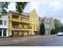 Straßenansicht mit mehrstöckigen Wohn- und Geschäftshäusern, darunter ein gelbes Gebäude mit einer Buchhandlung im Erdgeschoss und bepflanzten Balkonen. Daneben befinden sich weitere Häuser mit grüner und weißer Fassade, begleitet von Bäumen und Gehwegen.