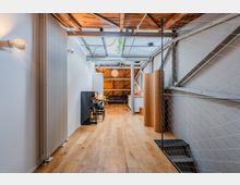 Langer, schmaler Innenraum im Loft-Stil mit hellem Holzboden, weiß gestrichenen Wänden und sichtbarer Holzbalkendecke unter einem Oberlicht. Rechts begrenzt ein Drahtnetz eine Treppe, links stehen ein hoher Heizkörper und ein Klavier; im Hintergrund sind eine kleine Küchenzeile und ein Sitzbereich zu sehen.
