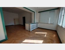 Ein leerer Innenraum mit Parkettboden, großen Fenstern mit Jalousien und einer weiß gestrichenen Heizkörperverkleidung. Das Zimmer ist durch einen offenen Durchgang mit einem angrenzenden weiteren Raum verbunden.