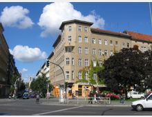 Straßenecke mit einem mehrstöckigen Wohn- und Geschäftshaus mit beiger Fassade und roten Ziegeldächern, teils bewachsen von Kletterpflanzen. Im Vordergrund sind Fußgänger, Radfahrer, parkende Autos und eine Ampel zu sehen, mit blauem Himmel und weißen Wolken im Hintergrund.