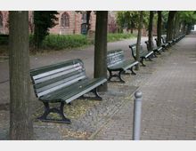 Eine gepflasterte Allee mit einer Reihe grüner Parkbänke entlang eines Gehwegs, gesäumt von Bäumen. Im Hintergrund ist eine gemauerte Fassade eines Backsteingebäudes mit gotischen Fenstern zu sehen.
