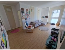 Wohnzimmer mit heller Einrichtung, bestehend aus einem grauen Sofa, einem weißen Sessel und einem Einbauschrank mit Regalfächern. Der Boden ist mit Holzfliesen ausgelegt, an den Wänden hängen Kinderzeichnungen und Kunstwerke, und das Zimmer hat ein großes Fenster mit halbtransparentem Rollo.