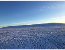 Schneebedeckte Landschaft mit leicht gewölbtem Hügel im Hintergrund, der von einem Holzzaun begrenzt wird. Zwei Menschen gehen in der Ferne unter einem klaren Himmel bei tiefstehender Sonne.