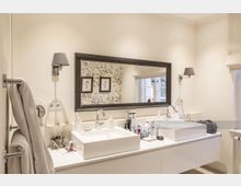 Modernes Badezimmer mit zwei weißen Waschbecken, einem breiten rechteckigen Spiegel in dunkel gerahmter Optik und beige gestrichenen Wänden. Rechts befindet sich ein Fenster mit heller Stoffjalousie, links hängen Handtücher an einem Wandhalter.