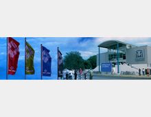 Moderne Veranstaltungsgebäude mit einem geschwungenen Dach und großen Glasfenstern; davor stehen Menschen in Gruppen sowie mehrere Fahrradständer. Links im Bild wehen bunte Fahnen mit Markenlogos, im Hintergrund sind Bäume zu sehen.