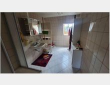 Ein Badezimmer mit hellen Fliesen an den Wänden und dem Boden. Zu sehen sind ein Waschbecken mit Spiegel, ein Handtuchheizkörper, ein kleines Fenster, eine Duschkabine sowie ein Schrank und Regal auf der rechten Seite.