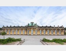 Ansicht des Schlosses Sanssouci in Potsdam, einem gelben Rokoko-Gebäude mit einer zentralen grünen Kuppel, großen Rundbogenfenstern und Stuckverzierungen. Vor dem Schloss befindet sich ein Kiesplatz und niedriges Grün.