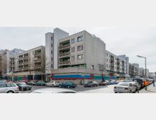 Wohngebäude in einer städtischen Umgebung, geprägt von moderner Betonarchitektur mit farbigen geometrischen Streifen an der Fassade. Im Vordergrund sind parkende Autos entlang einer gepflasterten Straße und kahle Bäume sichtbar.