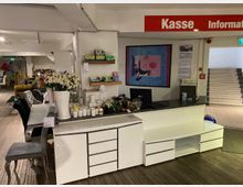 Kasse- und Informationsbereich in einem Möbelhaus mit weißem Tresen, auf dem Dekorationsartikel wie Blumen in einer Vase und Kerzen stehen. Im Hintergrund sind Möbel und ein Treppenaufgang zu sehen; ein rotes Schild mit der Aufschrift „Kasse Information“ hängt darüber.