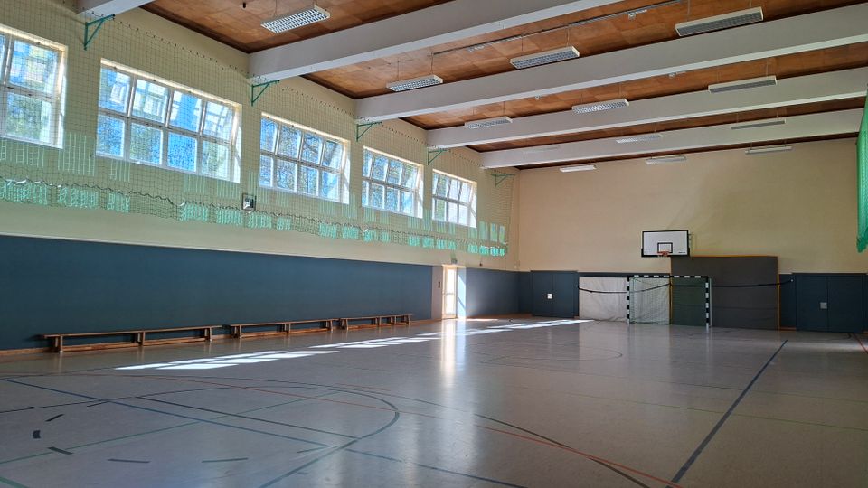 Große Sporthalle mit markiertem Hallenboden, langen Holzleistenbänken an der Wand und hohen Fensterreihen, die viel Tageslicht hereinlassen. An der Stirnseite sind ein Handballtor und ein Basketballkorb zu sehen, unter einer Decke mit Holzbalken und Deckenleuchten.