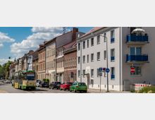 Straßenszene mit zweigeschossigen Wohnhäusern, teils mit Backsteinfassaden und Balkonen. Eine Straßenbahn der Linie 6 fährt an parkenden Autos vorbei, bei klarem Himmel mit einigen Wolken.