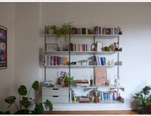 Ein helles Wohnzimmer mit einem weißen Wandsystem aus Regalen und Schubladen, gefüllt mit Büchern, Pflanzen, Dekorationsgegenständen und einer kleinen Lampe. Auf der linken Seite ist ein Poster an der Wand angebracht, und im Vordergrund stehen mehrere Zimmerpflanzen auf einem Holzboden.