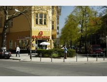 Straßenecke mit einer gelb gestrichenen Apotheke im Erdgeschoss eines Wohngebäudes, umgeben von Gehwegen, parkenden Autos und Bäumen entlang der Straße. Im Vordergrund kleine Büsche und vereinzelte Fußgänger.