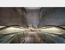 Treppen- und Rolltreppenabgang in einer modernen U-Bahn-Station mit grauen Steinwänden und Glasgeländern. Der Bereich ist hell beleuchtet, und am unteren Ende des Abgangs ist ein Bodenbereich mit grünlichen Akzenten zu sehen.